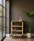 A rustic wooden wine rack cart with casters, styled in a modern room next to a large potted plant and a window.