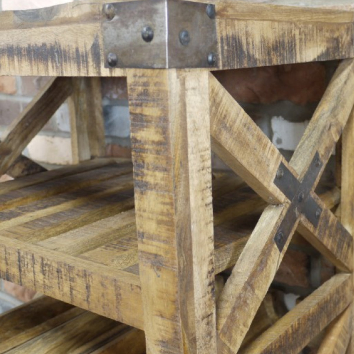 Rustic wooden side table with distressed natural wood finish, dark metal hardware, and X-brace support on a brick background.
