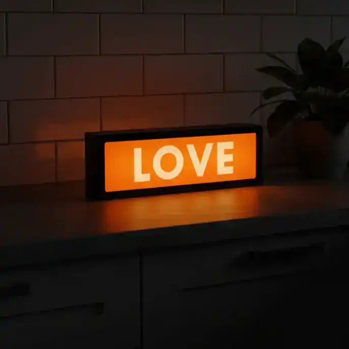 Orange LED sign with 'LOVE' text on a dark surface with a plant in the background.