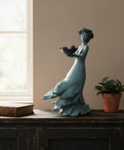 Decorative statue of a woman holding birds on a wooden surface with a plant and book in the background.