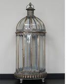 An ornate vintage-style birdcage lantern with a silver-toned metal frame, clear glass panels, and a decorative finial on top, photographed against a plain light gray wall.