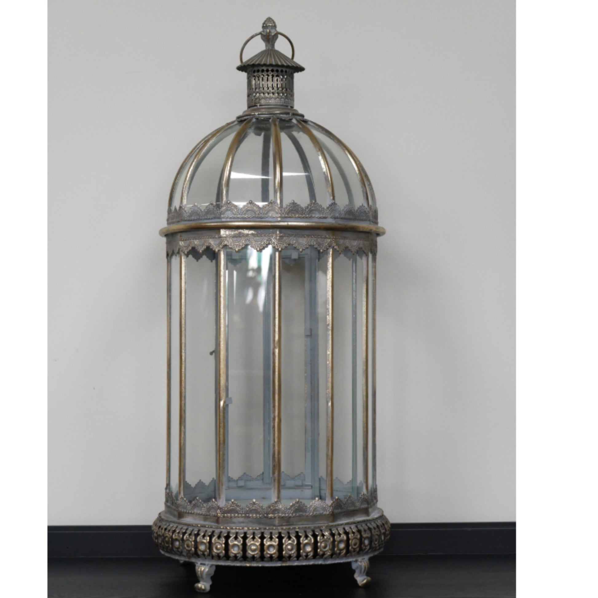 An ornate vintage-style birdcage lantern with a silver-toned metal frame, clear glass panels, and a decorative finial on top, photographed against a plain light gray wall.