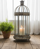 A vintage-style metal birdcage lantern with a lit white pillar candle inside, placed on a rustic wooden floor in front of sheer white curtains. A small green potted plant sits to the left.