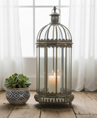 A tall vintage-style metal birdcage lantern with a lit white pillar candle inside, placed on a rustic wooden floor next to a small potted succulent in front of sheer white curtains.