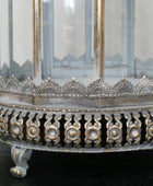 Close-up of an ornate silver-toned metal birdcage or lantern base with decorative filigree, clear glass panels, and a small claw-foot stand on a dark surface.