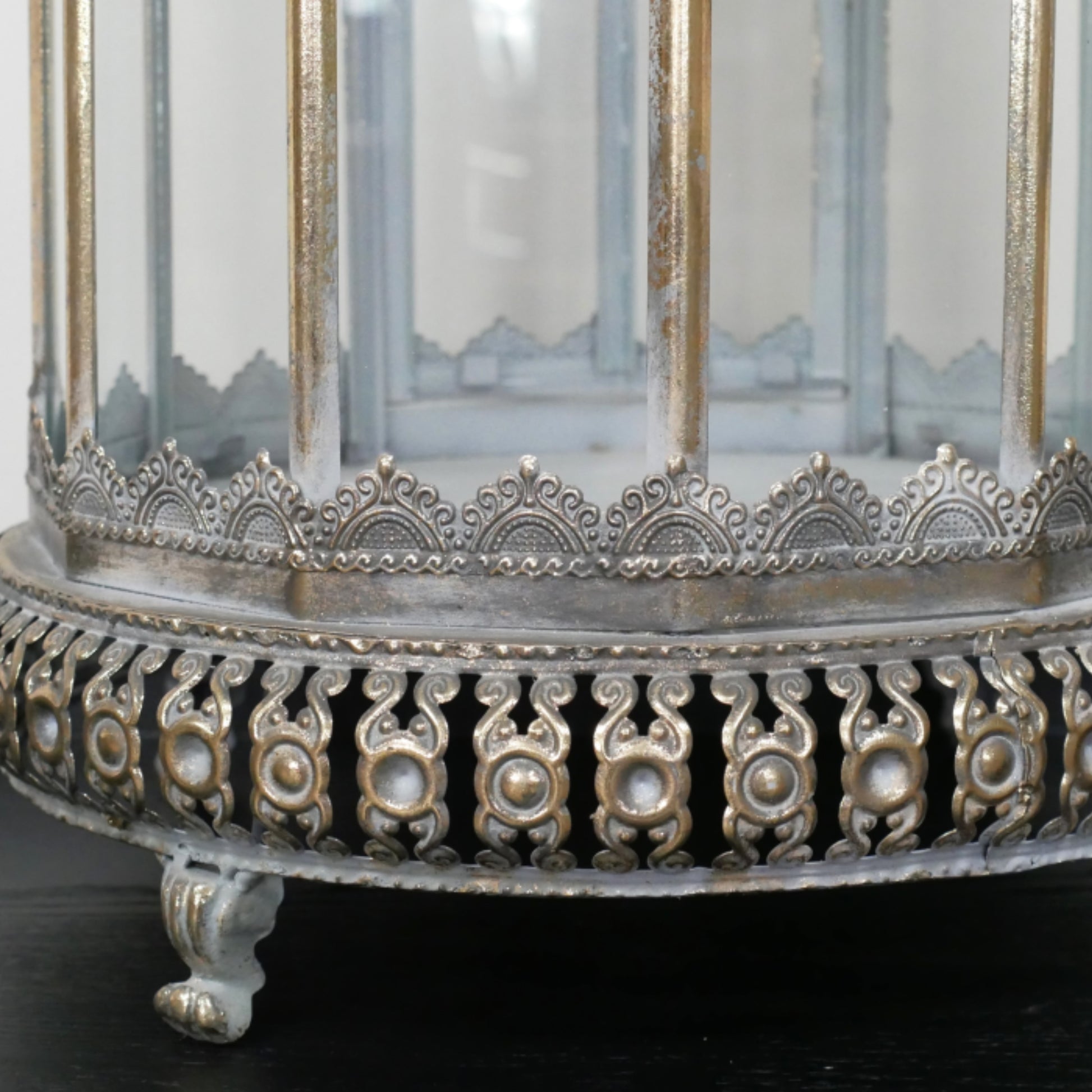Close-up of an ornate silver-toned metal birdcage or lantern base with decorative filigree, clear glass panels, and a small claw-foot stand on a dark surface.