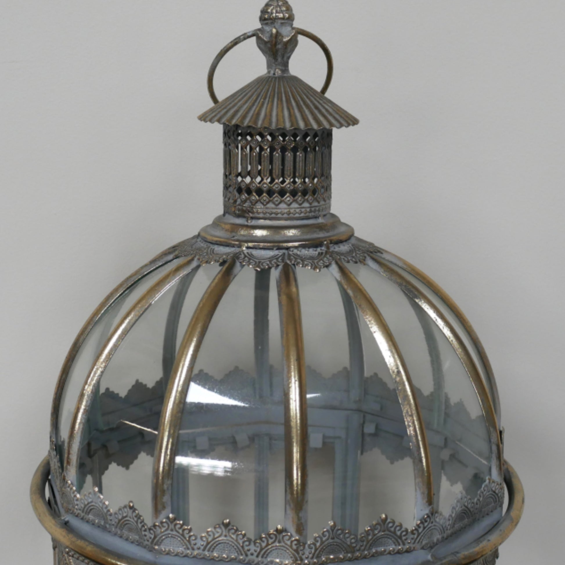 A close-up of an ornate vintage lantern with a clear ribbed glass dome, silver-toned metal frame, and decorative top finial against a neutral gray background.
