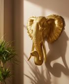 Gold elephant head wall decoration on a beige wall with a plant in the foreground.