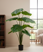 A large artificial monstera plant in a black pot, styled in a bright, modern room next to a bookshelf and window.