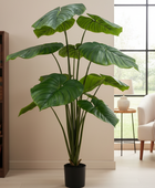 A large artificial monstera plant with glossy green leaves in a black pot, placed in a modern living room setting next to a bookshelf and window.