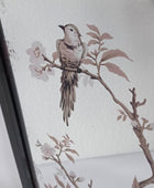 A close-up of a framed wall art print featuring a small bird perched on a branch with pale blossoms, shown against a white textured background.