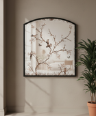 Arched wall mirror with a black frame and a delicate floral branch print, hanging on a beige wall next to a potted plant.