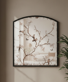 Arched decorative wall mirror with a black frame and a delicate branch-and-bird print, mounted on a beige wall next to a green plant.