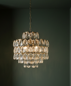 A tiered crystal chandelier with clear teardrop-shaped glass drops and a gold-toned metal frame, hanging against a dark green wall.
