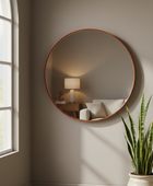 A large round wall mirror with a warm copper-toned frame hangs on a taupe-gray wall in a softly lit room. The mirror reflects a lamp and sofa, and a potted snake plant sits in the lower right corner.