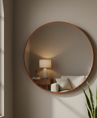A large round wall mirror with a warm copper-toned frame hanging on a taupe-gray wall in a styled room. The mirror reflects a softly lit interior with a table lamp, side table, and sofa, while a green snake plant is visible in the lower right corner.