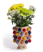 A beige ceramic vase covered in colorful glossy spherical bumps, filled with yellow, green, and white daisies, photographed on a white background.