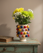 A cream-colored ceramic vase decorated with colorful glossy pom-pom beads, filled with yellow and white flowers, set on a distressed teal wooden table next to an old book against a beige wall.