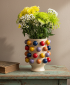 A cream-colored ceramic vase covered in colorful glossy pom-pom details, filled with yellow, green, and white flowers, sitting on a distressed teal wooden table next to an old book against a beige wall.