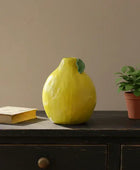 Yellow ceramic vase on a wooden surface with a book and potted plant in the background.