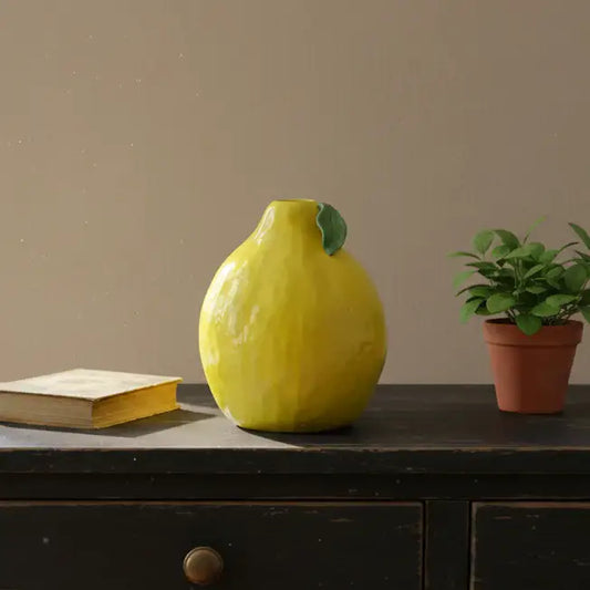 Yellow ceramic vase on a wooden surface with a book and potted plant in the background.
