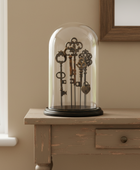 A set of ornate antique-style metal keys displayed inside a clear glass cloche dome on a round black base, set on a distressed wooden console table in a warm beige room.