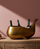 Gold whale-shaped wine cooler with bottles on a wooden surface against a pink wall.