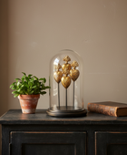 Decorative gold heart sculpture under a glass dome on a wooden surface with a plant and book in the background.