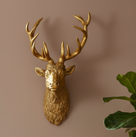 Gold deer head wall decoration on a brown wall with a plant in the corner.