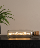 A clear glass bottle with a cork stopper lies on a dark wooden surface, lit with warm glowing filament text that reads 'Home Sweet Home'. A potted green fern sits to the left against a plain beige wall.