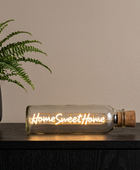 A clear glass bottle-style light with a cork stopper lies on a dark wooden surface, glowing with warm light and the words “Home Sweet Home” in script. A potted fern and beige wall are visible in the background.