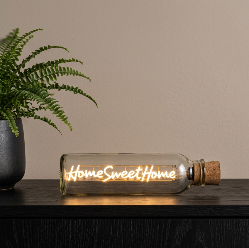 A clear glass bottle-style light with a cork stopper lies on a dark wooden surface, glowing with warm light and the words “Home Sweet Home” in script. A potted fern and beige wall are visible in the background.