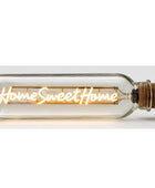 A clear glass bottle-style light with a cork stopper, glowing with warm yellow light and the words 'Home Sweet Home' across the front, shown on a plain light background.