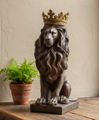 A bronze-finished lion statue wearing a gold crown, displayed on a rustic wooden surface next to a small potted fern against a textured beige wall.