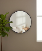 A round wall mirror with a thin dark frame hangs on a warm taupe wall, reflecting a softly lit interior. A potted green plant is visible on the left, and a bright window is on the right.