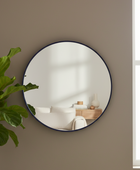 Round wall mirror with a thin dark navy frame, mounted on a warm taupe wall next to a green fiddle-leaf fig plant. The mirror reflects a softly lit modern interior with white furniture and a wooden side table.