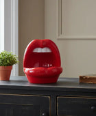 Red lip-shaped object on a wooden surface with a plant and book in the background.