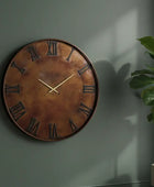 Bronze wall clock with Roman numerals on a dark green wall with a plant to the right.