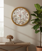 Round wall clock with a gold frame and a vintage-style bird and floral illustration on the face, styled on a beige wall above a rustic wooden bench with a potted plant nearby.
