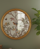 Round framed wall mirror with an ornate gold frame and a vintage-style botanical illustration of a bird and flowers, mounted on a sage green wall next to a potted fiddle-leaf fig plant.