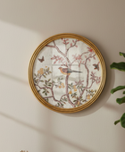 Round framed wall art featuring a vintage-style bird and floral branch illustration on a textured off-white background, with a gold frame, mounted on a beige wall next to a green plant.