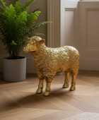 A metallic gold sheep figurine standing on a light wood herringbone floor next to a potted green fern in a gray pot, set against a beige paneled wall.