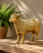 A metallic gold sheep figurine stands on a rustic wooden table beside a potted green fern, with warm sunlight casting shadows on a beige wall.