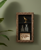 A decorative wall-mounted cabinet with an ornate bronze frame, three shelves, and display items including a gold bird figurine, two glass bottles, and a framed fern print, shown against a muted green wall with plant leaves at the left edge.
