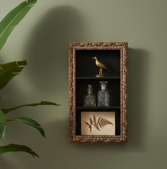A decorative wall-mounted cabinet with an ornate bronze frame, three shelves, and display items including a gold bird figurine, two glass bottles, and a framed fern print, shown against a muted green wall with plant leaves at the left edge.