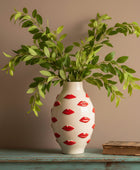 An off-white ceramic vase with a repeating red lips pattern, filled with dried brown leaves, styled on a white marble surface next to a red candlestick holder.