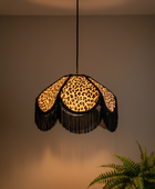 A hanging pendant light with a black rod and canopy, featuring a leopard-print shade with black fringe, shown lit against a warm beige wall with a green plant in the lower right corner.
