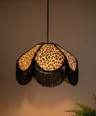 A hanging pendant lamp with a leopard-print fabric shade and long black fringe, shown lit against a warm neutral wall with a small plant visible in the lower right corner.