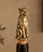 A metallic gold-toned leopard bottle stopper with cutout details, mounted on a cork and dark glass bottle against a warm beige wall background.