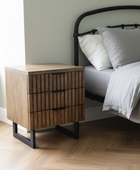 Wooden nightstand next to a bed with gray bedding in a bedroom setting.